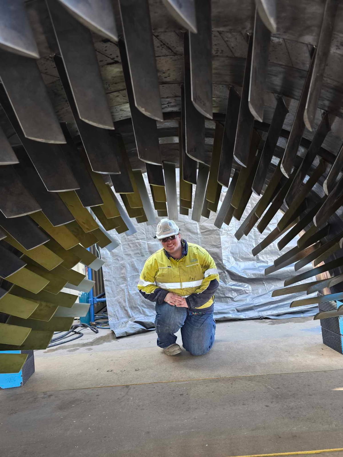 Beth, con casco y chaleco de seguridad, se arrodilla debajo de una máquina grande.