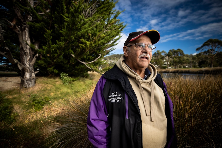 An Aboriginal man wearing a cap and jacket standing on the banks of a river.