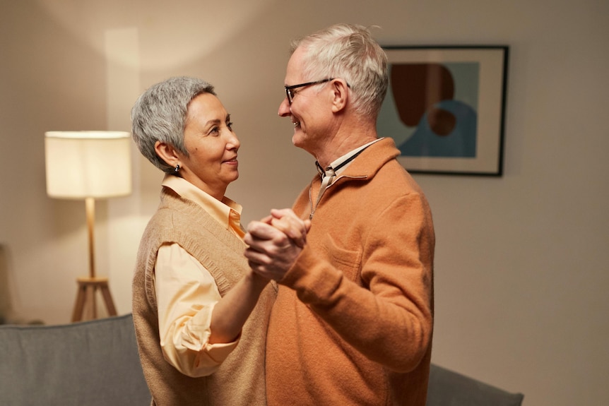an elderly man and woman dance together in their living room smiling, they both are wearing orange 