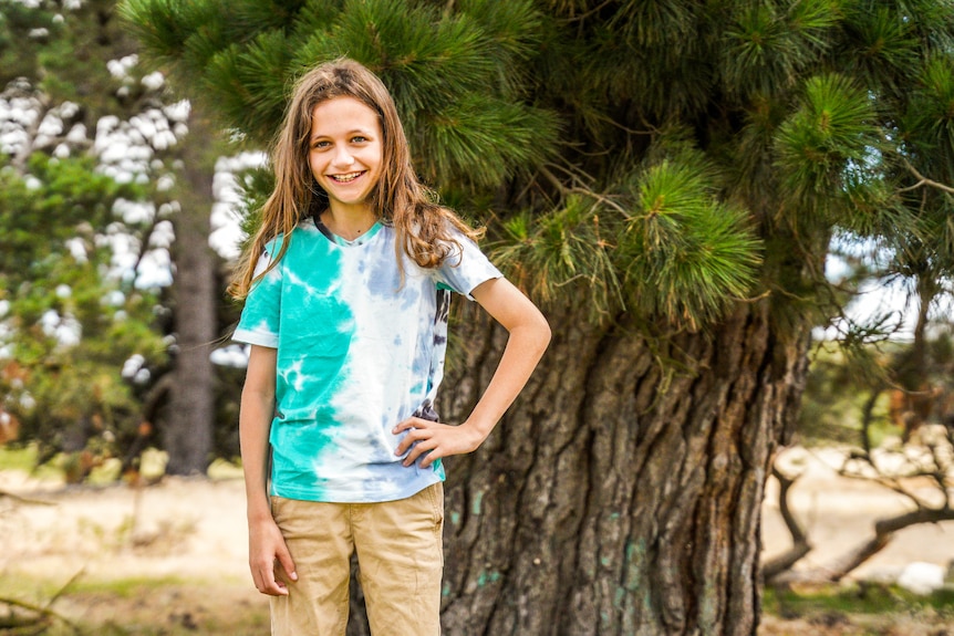 A boy stands on a tree log smiling with a hand on his hip.