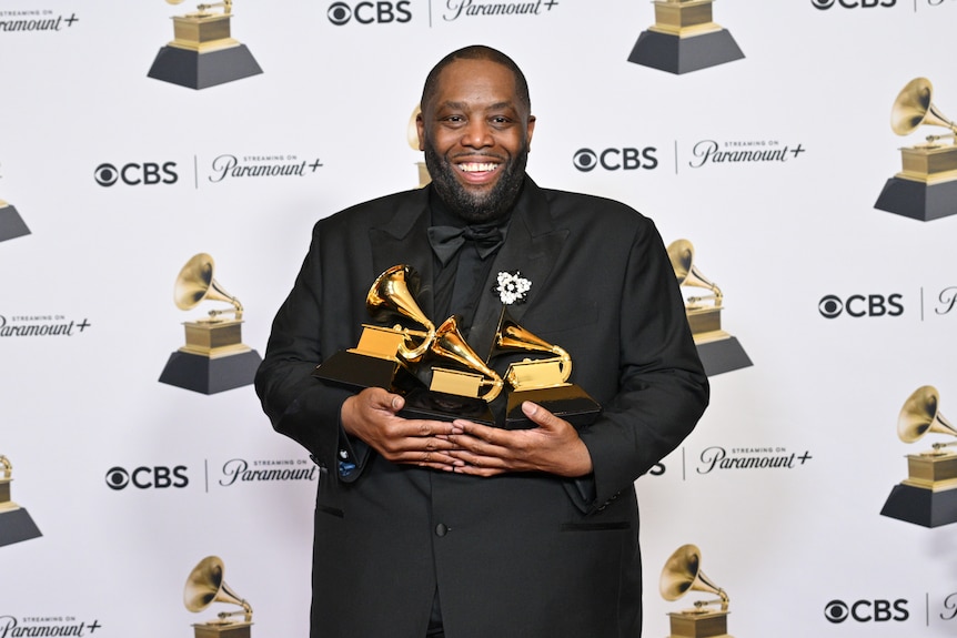 Killer Mike holding three Grammys and smiling widely, wearing all black, standing in press room with pics of Grammys and logos