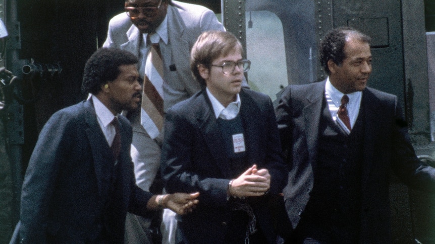 Several men in suits lead another man wearing glasses in handcuffs