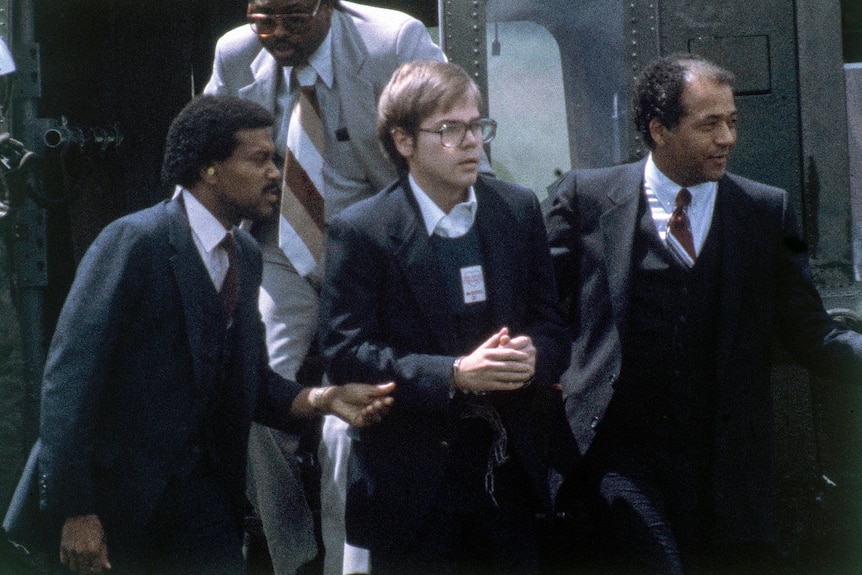 Several men in suits lead another man wearing glasses in handcuffs