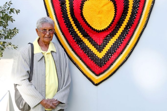 Aboriginal elder stands with large crocheted shield in colours of the Aboriginal flag