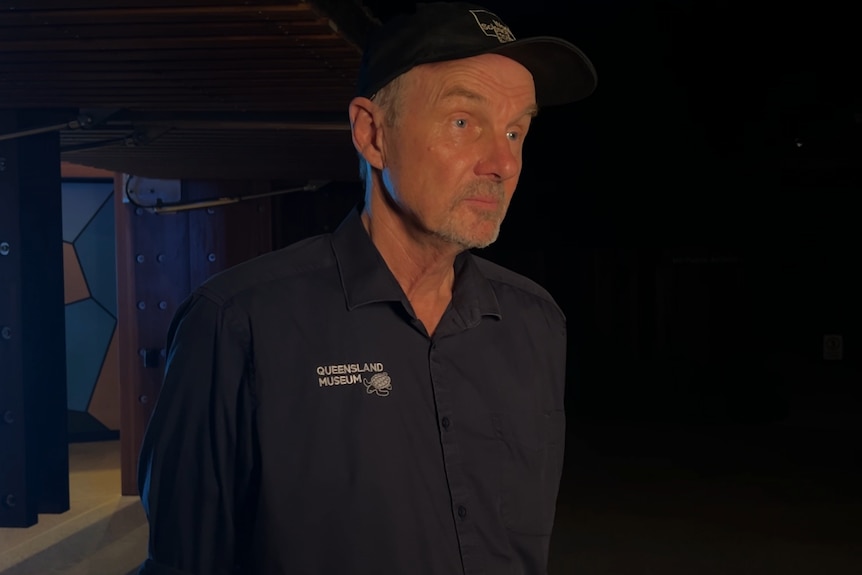 A man in a Queensland Museum shirt looks off camera, dark background