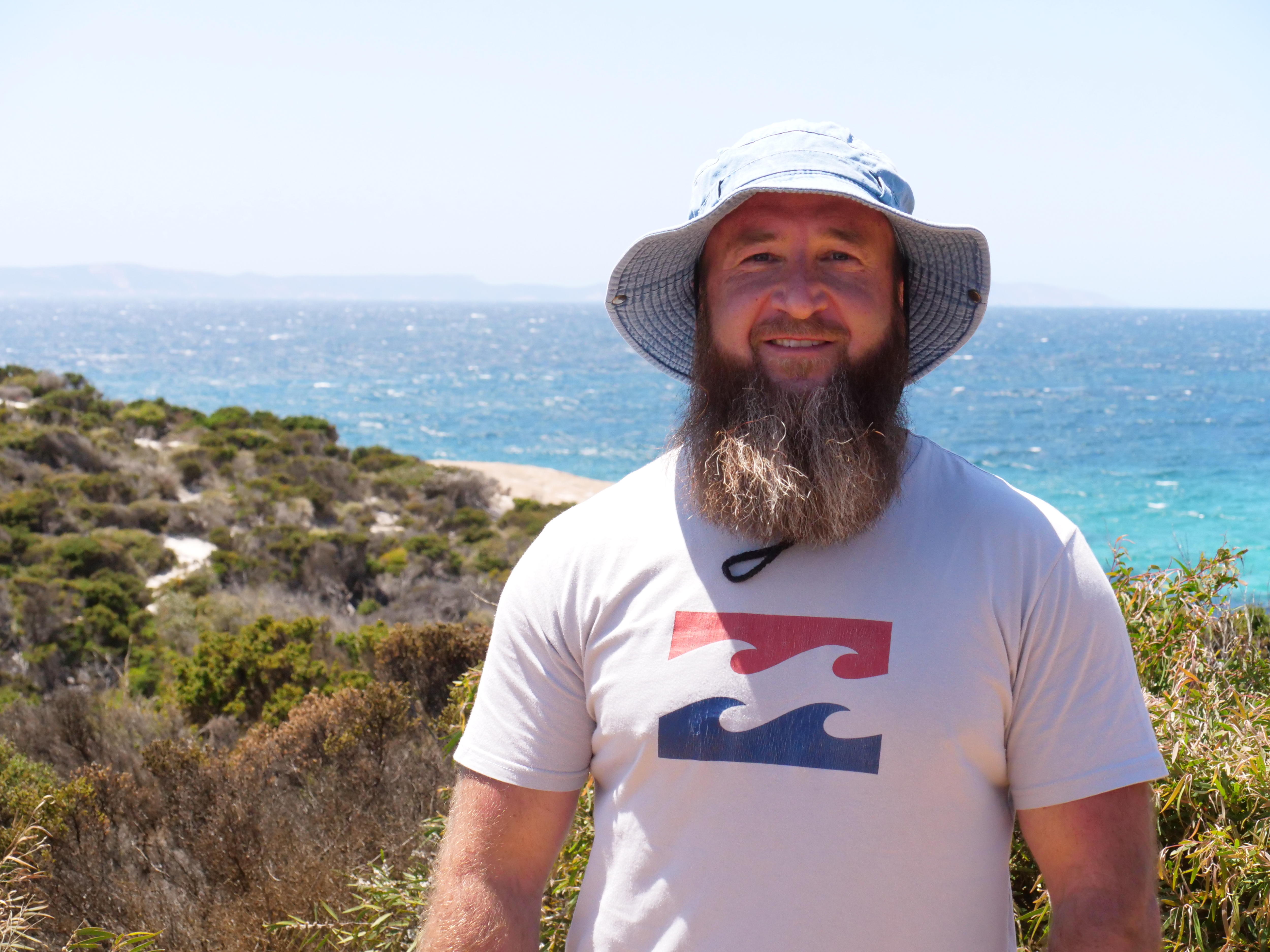 A smiling, bearded man in a slouch hat stands in front of the ocean.