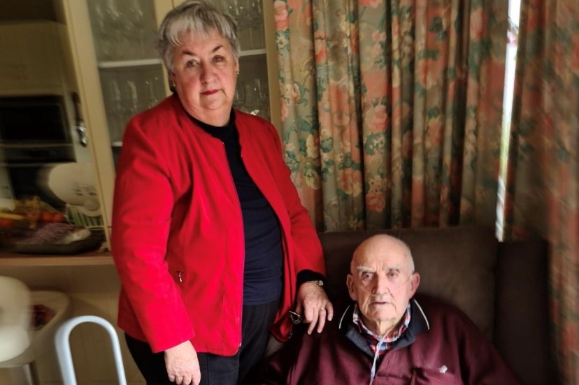 Older woman stands next to sick old man.