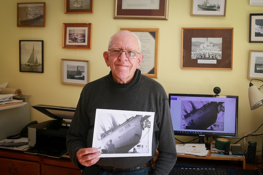 A man smiles at the camera holding a photo of a ship. There are framed photos of boats behind him.