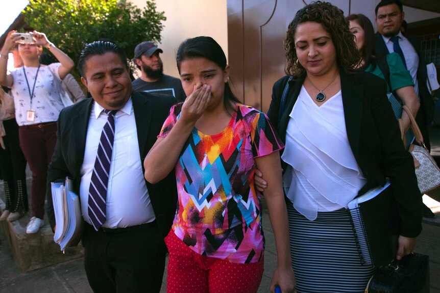 A young woman wipes her nose as she walks from court flanked by lawyers.