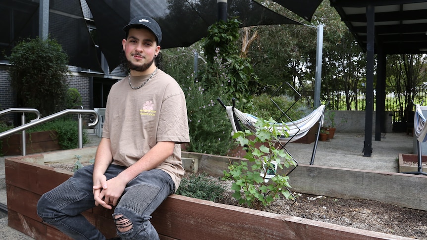 A young man in ripped jeans and a hat sits next to a garden