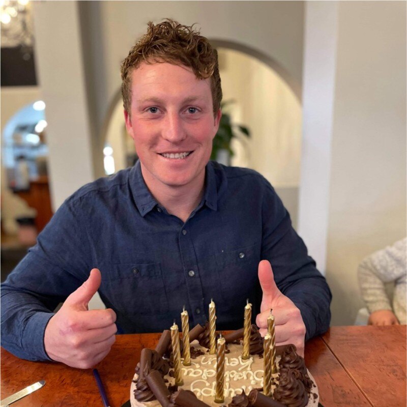 A smiling, ginger-haired man in a dark shirt sitting indoors.