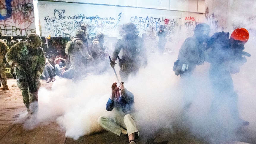 a civilian cowers on the ground as police in camouflage gear step around him through a cloud of tear gas.