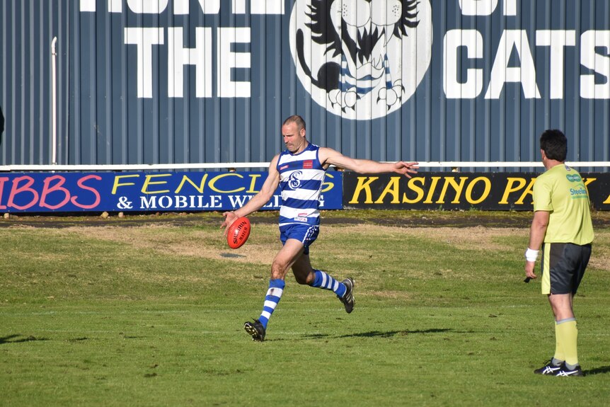 A man in a blue and white striped football uniform kicks a football, umpire standing next to him