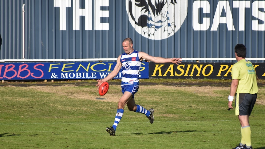 A man in a blue and white striped football jersey and blue shorts kicks a football, umpire standing next to him