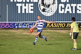A man in a blue and white striped football jersey and blue shorts kicks a football, umpire standing next to him