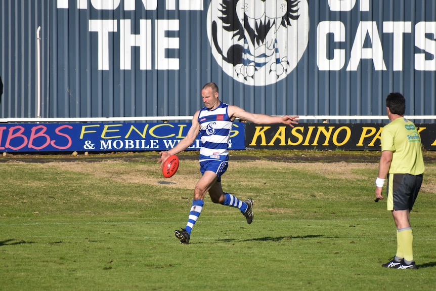 A man in a blue and white striped football uniform kicks a football, umpire standing next to him