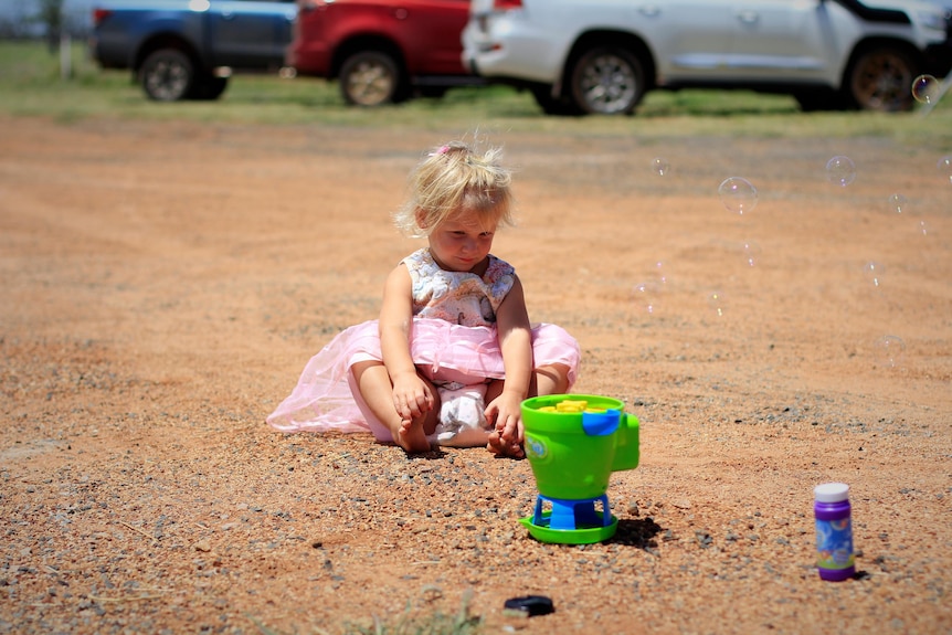 A two-year-old girl sits on red dirt watching a bubble machine
