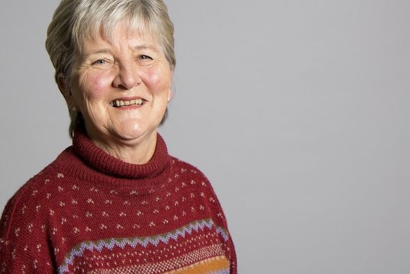 An older woman smiles at the camera against a grey background.