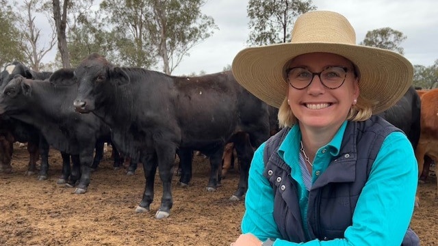 A woman in a large hat stands in front of cows in a paddock.