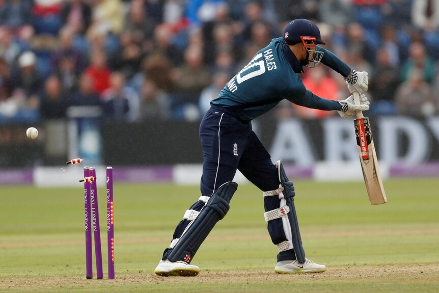 A cricketer wields a bat while a ball hits the wicket behind him