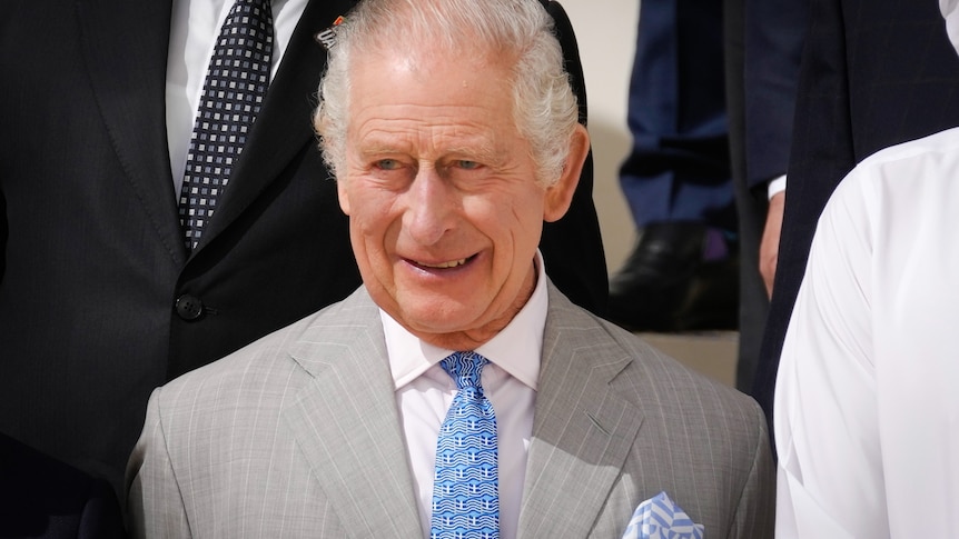 King Charles III smiles while wearing a grey suit with a blue tie with the Greek flag and a matching handkerchief