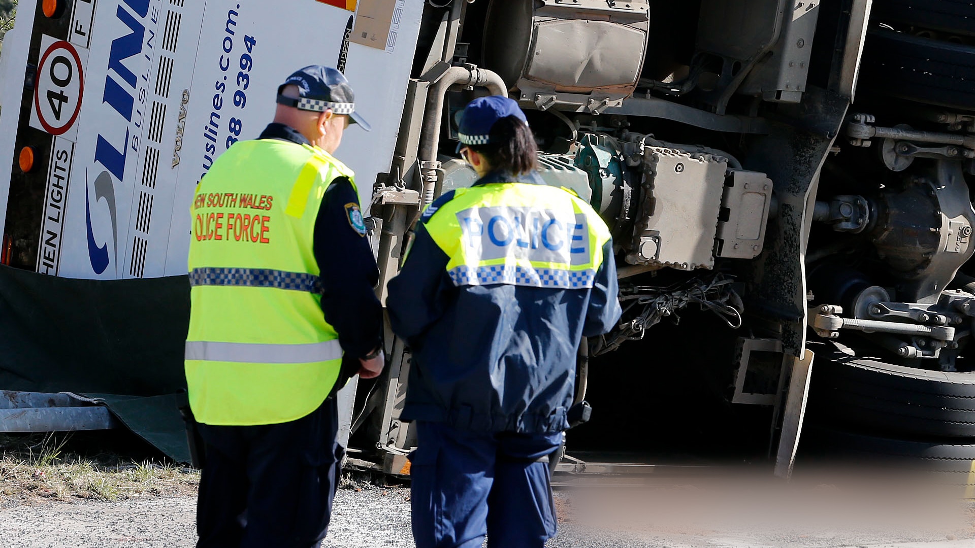 Two police officers with their backs turned stand in front of a bus that is turned on its side on a road.