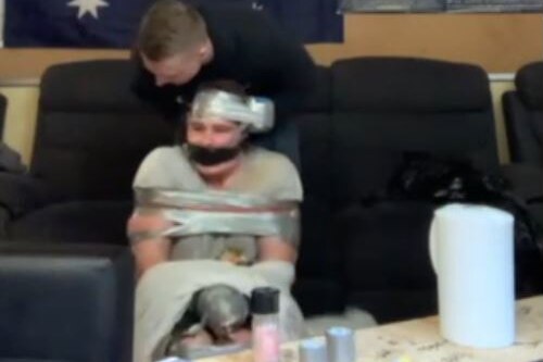 A man is bound and gagged by tape while another man stands up behind him.