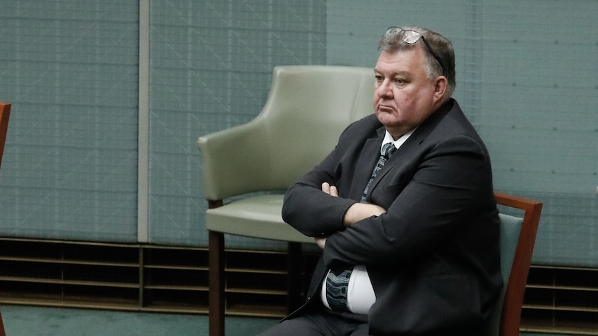 A white man with grey hair in a suit and tie sitting with his arms cross on a chair on the floor of parliament