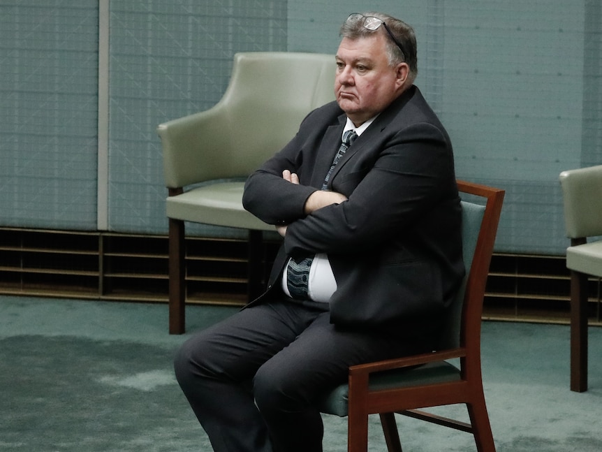 A white man with grey hair in a suit and tie sitting with his arms cross on a chair on the floor of parliament