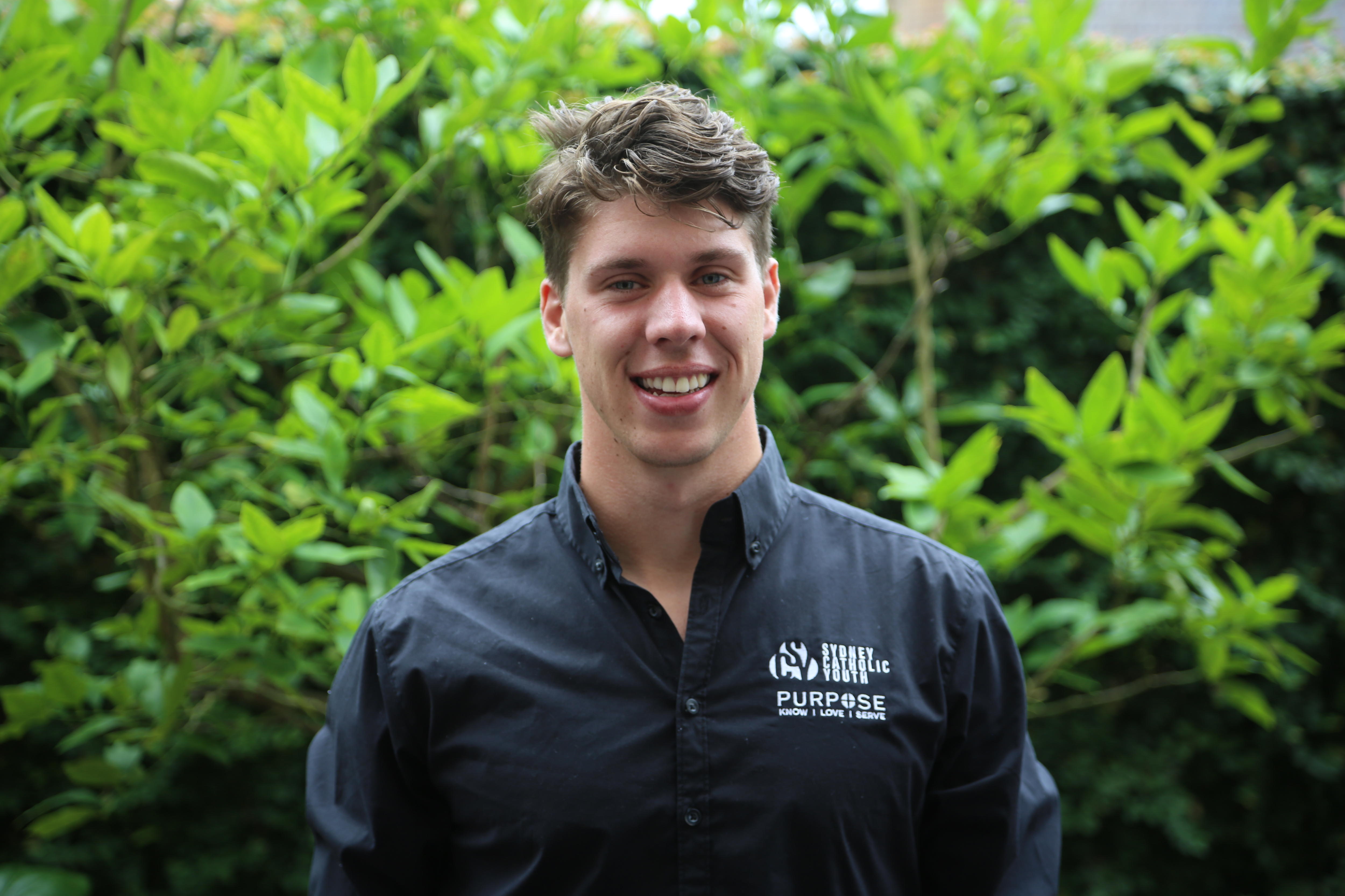 Young man wearing black shirt, standing in front of greenery.