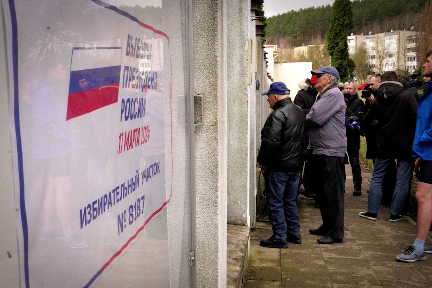 People standing in a line outdoors, near a sign with Russian characters on it.