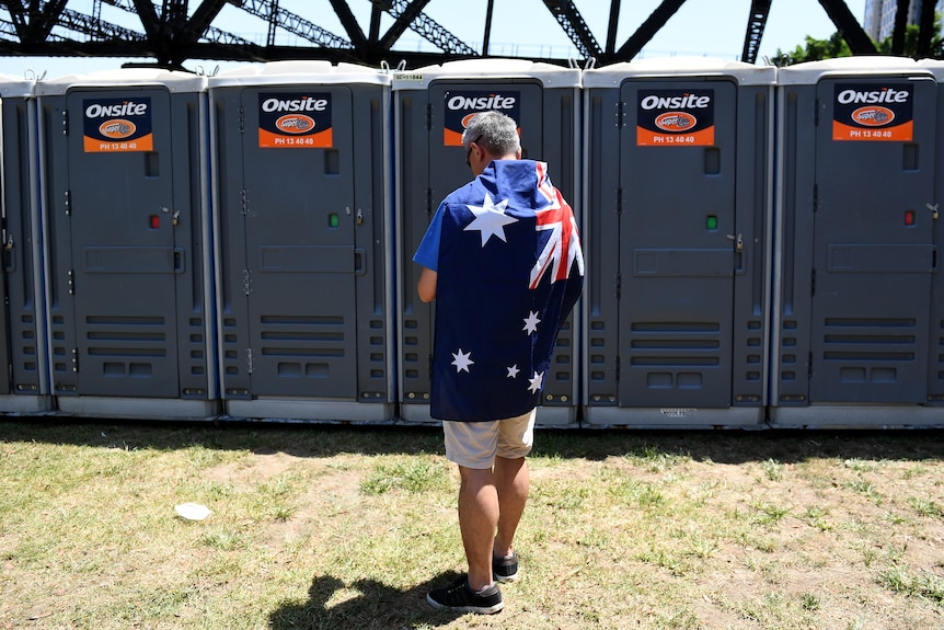 Grey-haired man wearing blue t-shirt, light shorts, an Australian flag draped over his shoulder waits at a row of portaloos.