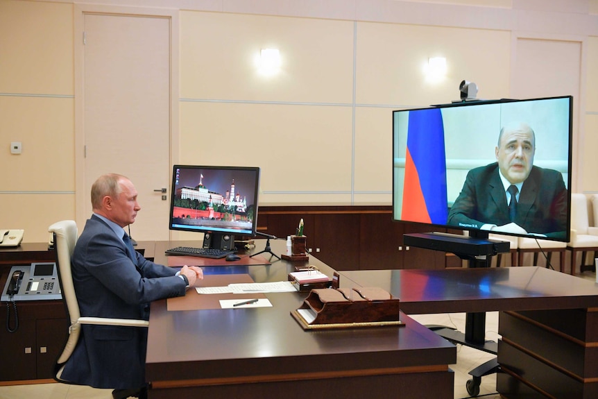 Russian President Vladimir Putin sits at a desk and listens to Mikhail Mishustin, displayed on TV screen on the right.