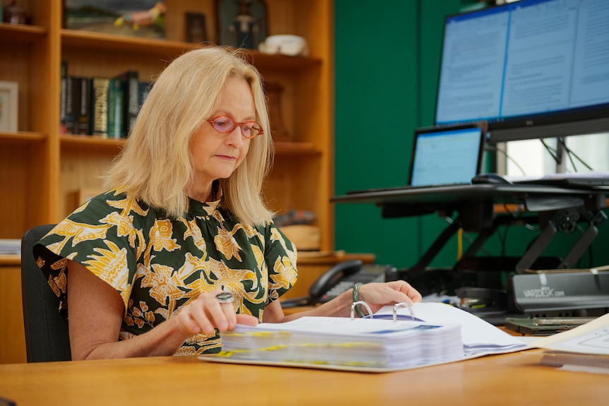 A woman sitting at her desk, with bookshelves in the background, and looking through a thick file of papers. 