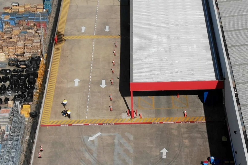 Two workers in high visibility workwear walk down the Amazon warehouse loading dock driveway pushing a trolley.