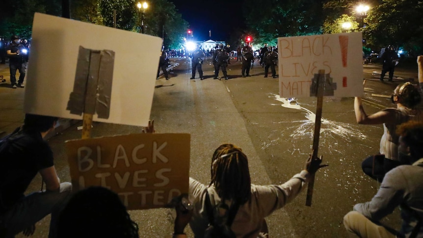 A group of protesters holding signs saying "Black Lives Matter" sit on a wide street, opposed by a line of police in riot gear.