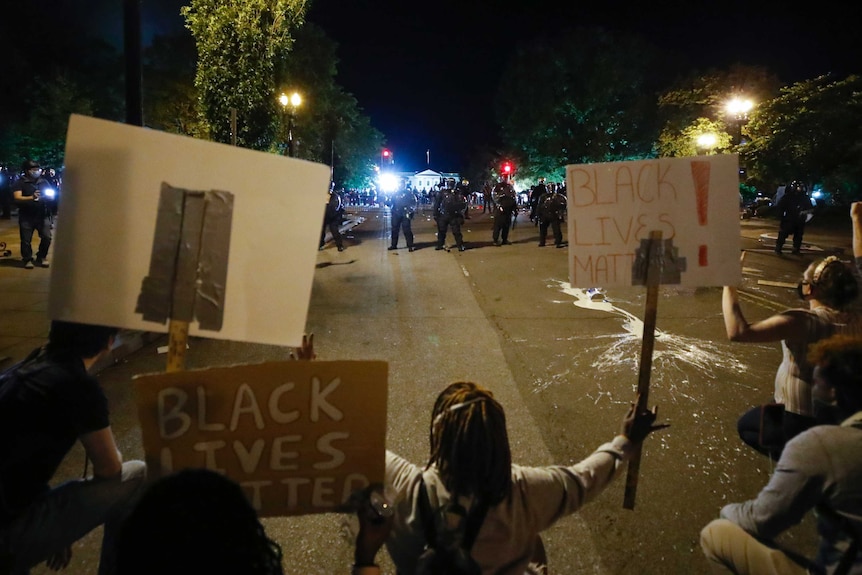 A group of protesters holding signs saying "Black Lives Matter" sit on a wide street, opposed by a line of police in riot gear.