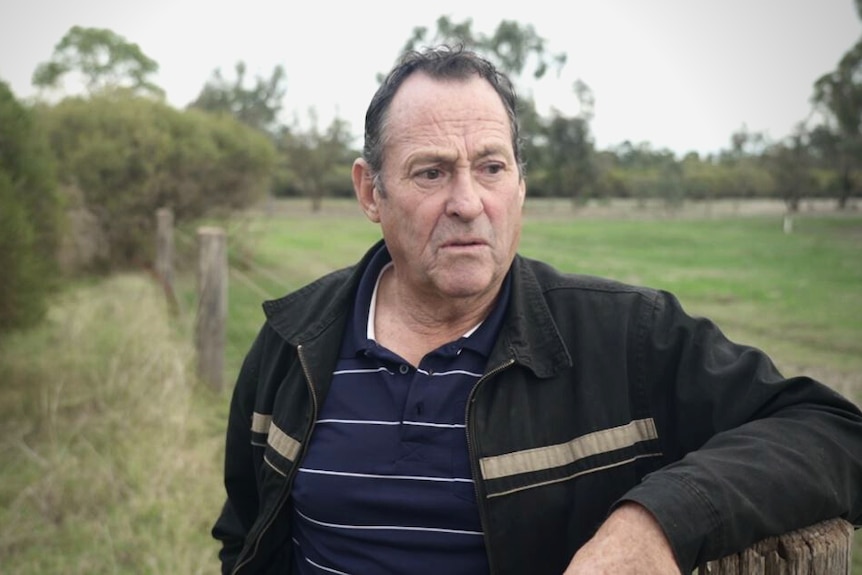A man in tshirt and casual jacket leans on fence on rural property while looking to distance