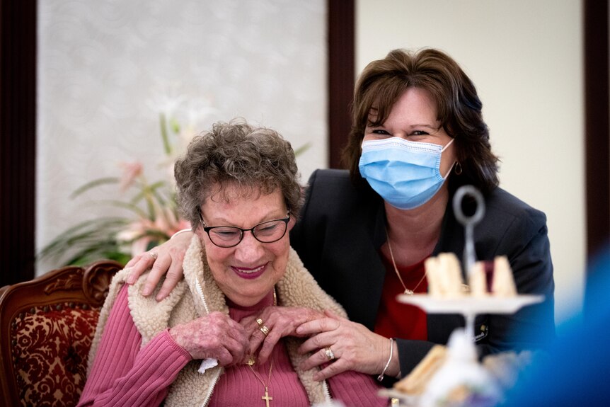 A woman with a mask on with her arm around an older woman, both laughing.