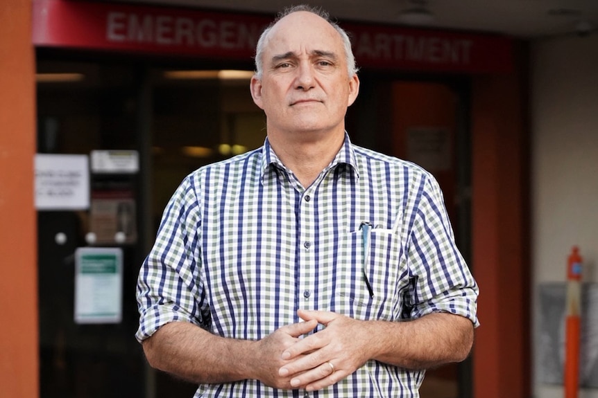 A bald man with a little bit of gray hair on side, wears white and blue shirt, posing for a photo outside emergency hospital.