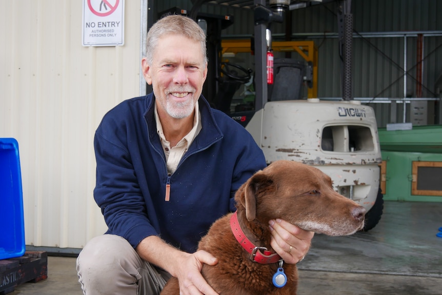 A man kneels next to a brown Labrador  in front of a shed. 