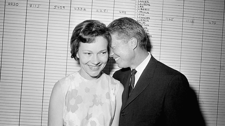 A black and white photo of Jimmy Carter leaning into his wife Rosalynn with a smile on their faces.