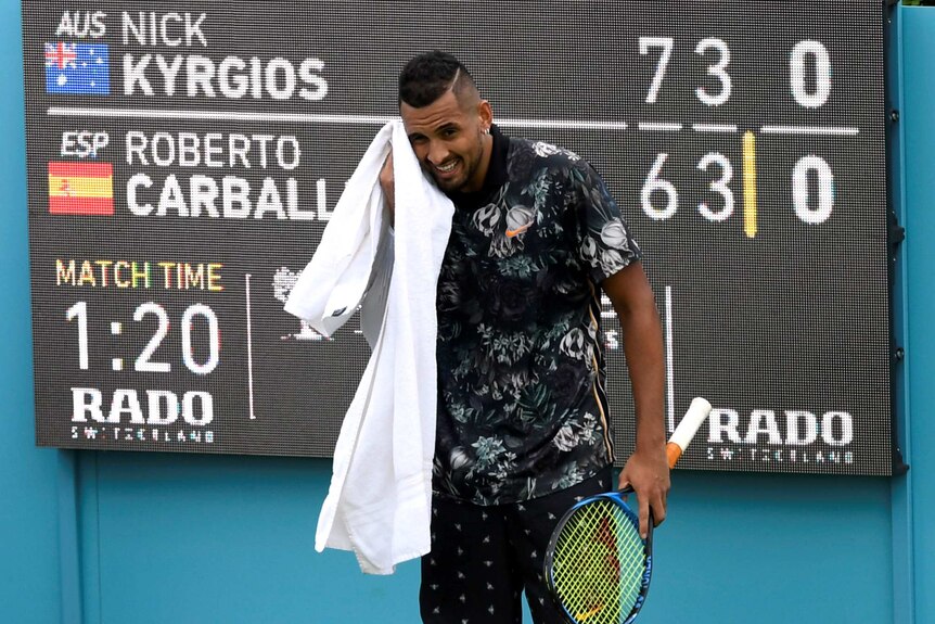 With the scoreboard in the background, Nick Kyrgios is seen grimacing as he wipes his face with a white towel.