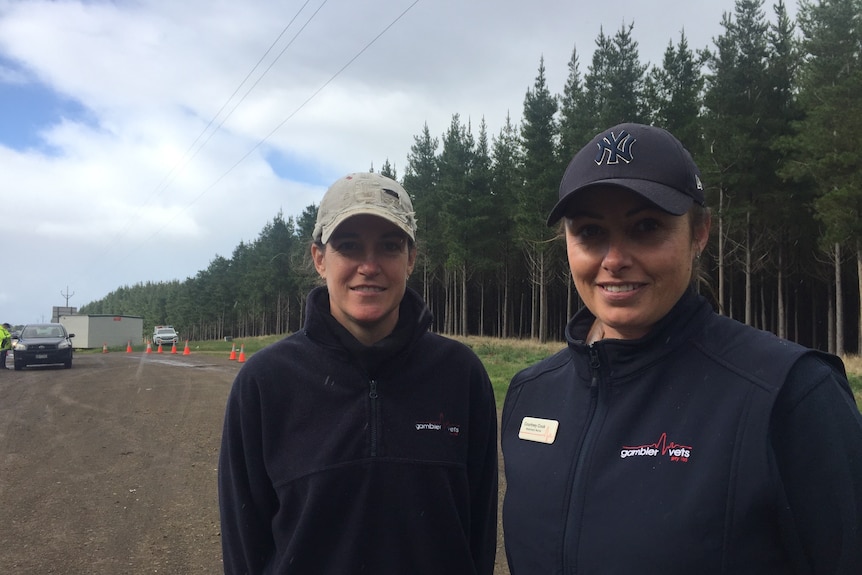 Two women wearing caps stand in front of trees near a border checkpoint