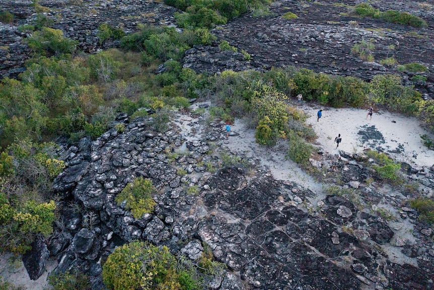 An aerial view of several people standing on a patch of white sand surrounded by rock formations and greenery. 