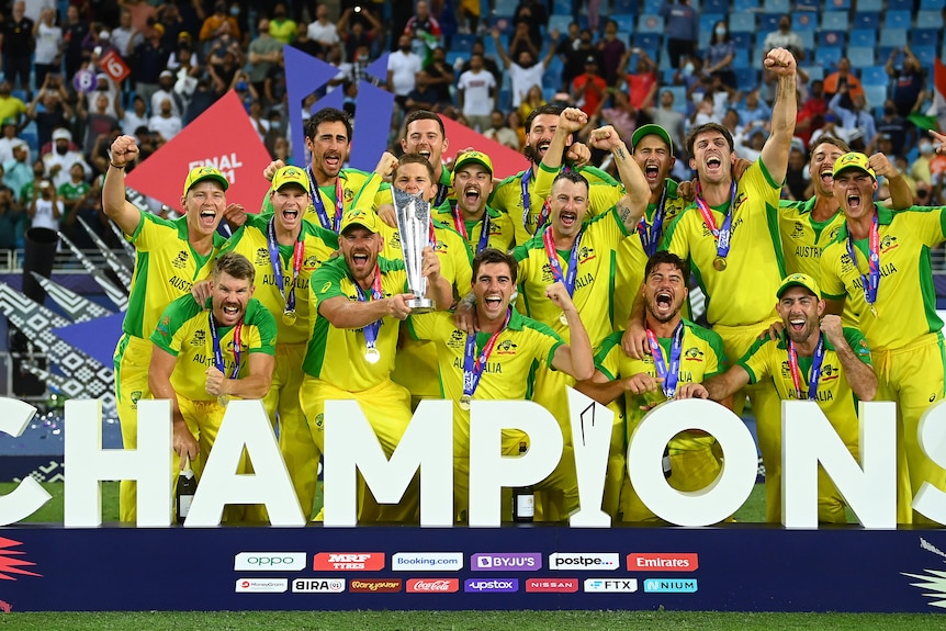 A group of Australian cricketers roar in triumph as they celebrate with the T20 World Cup trophy in front of a 'Champions' sign.