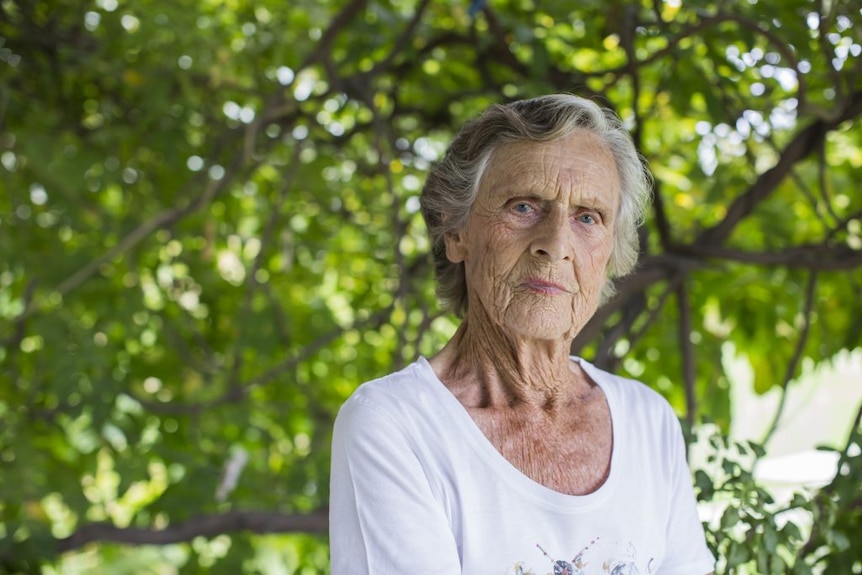 A grey haired woman stares to camera, standing in front of a background of lush tree leaves.