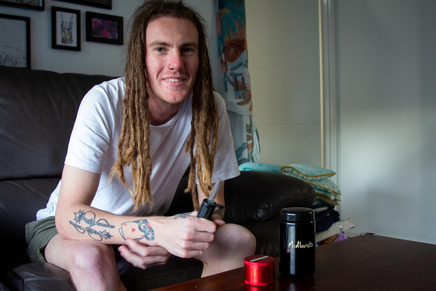 A man with dreadlocks and a white shirt sits on a couch, holding a vape with a cannabis grinder in front of him on a table. 