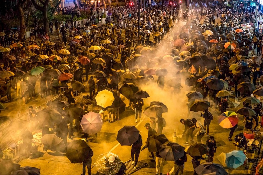 the mist of tear gas is spread over thousands of gathered protesters, many carry umbrellas.