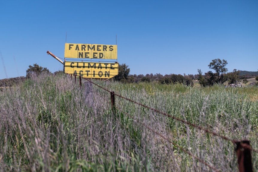 A yellow sign reading 'Farmers need climate action' in a paddock with barbed wire fence